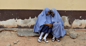 two veiled young girls smiling, sitting against a chipped wall
